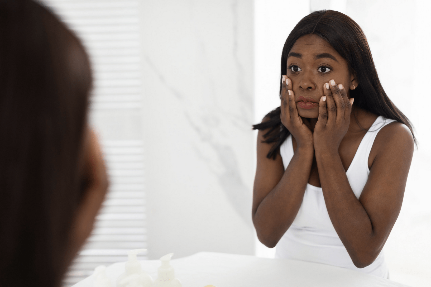 younger black woman with long black hair looking at her face in the mirror with hands on side of her face